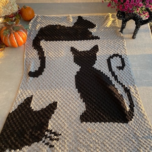 Op de afbeelding: Een grijs gehaakt deken met zwarte katten silhouetten. Het deken heeft drie katten in verschillende poses, waaronder een kat die opgerold ligt, een kat die zit en een kat die staat.