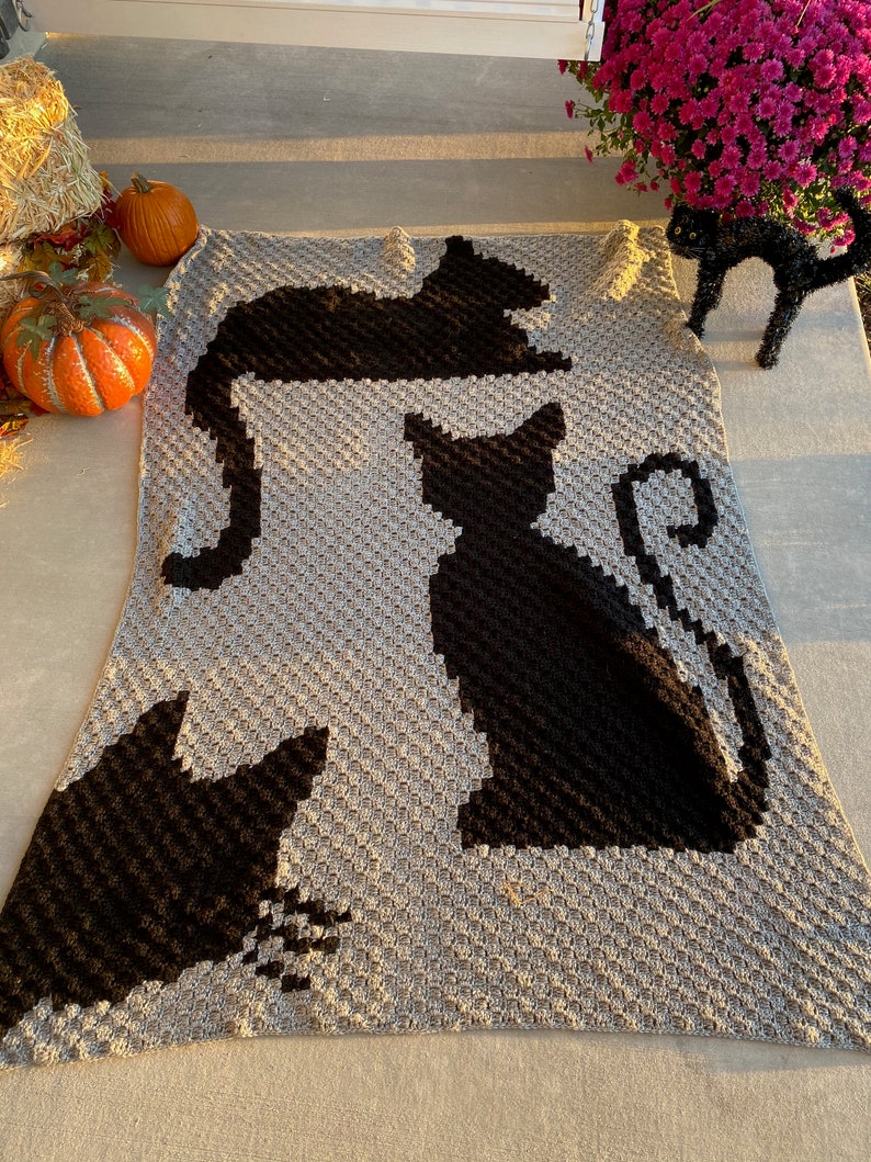 Op de afbeelding: Een grijs gehaakt deken met zwarte katten silhouetten. Het deken heeft drie katten in verschillende poses, waaronder een kat die opgerold ligt, een kat die zit en een kat die staat.