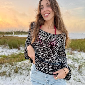 Könnte beinhalten: Eine Frau trägt ein schwarzes Häkeltop mit einem rosafarbenen Bikini-Oberteil darunter und hellblaue Jeans-Shorts. Sie steht an einem Strand mit Sand und Gras im Hintergrund.