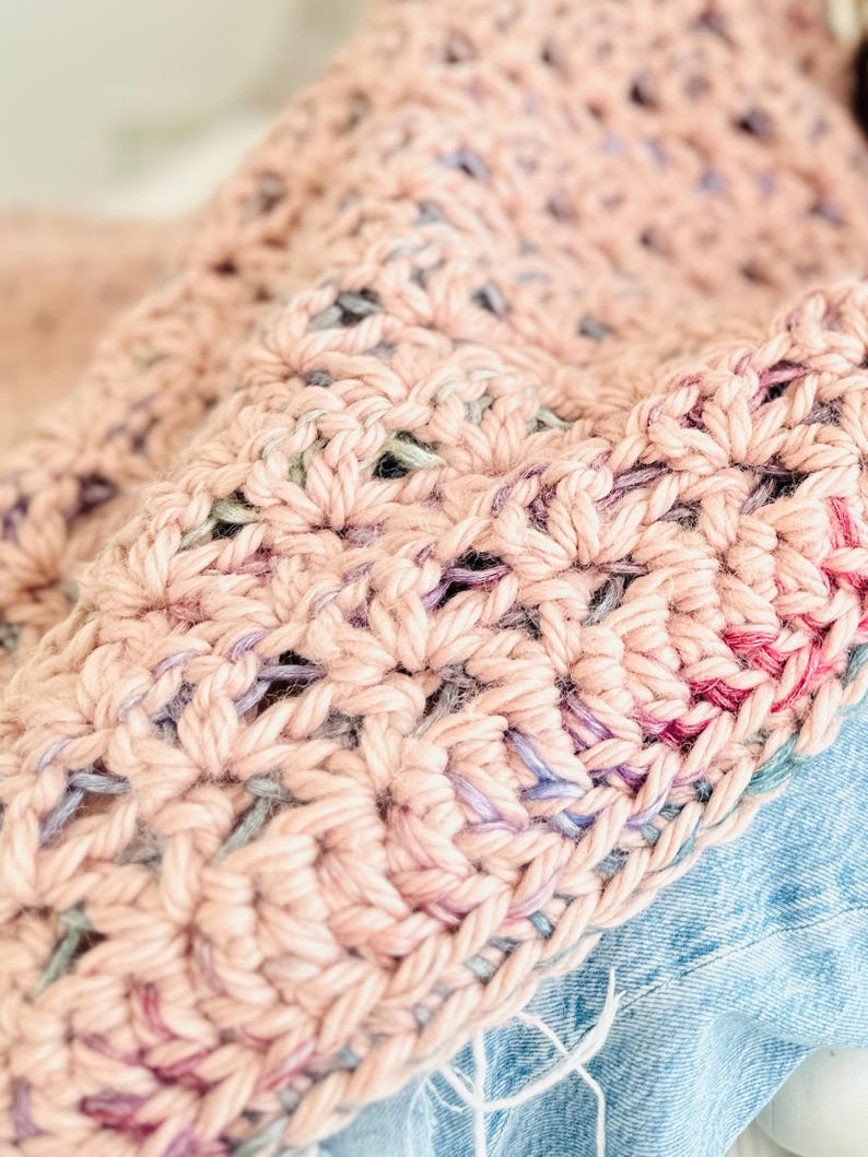 May include: A close-up of a pink crocheted blanket with a multi-colored yarn. The blanket is folded and draped over a blue denim surface.