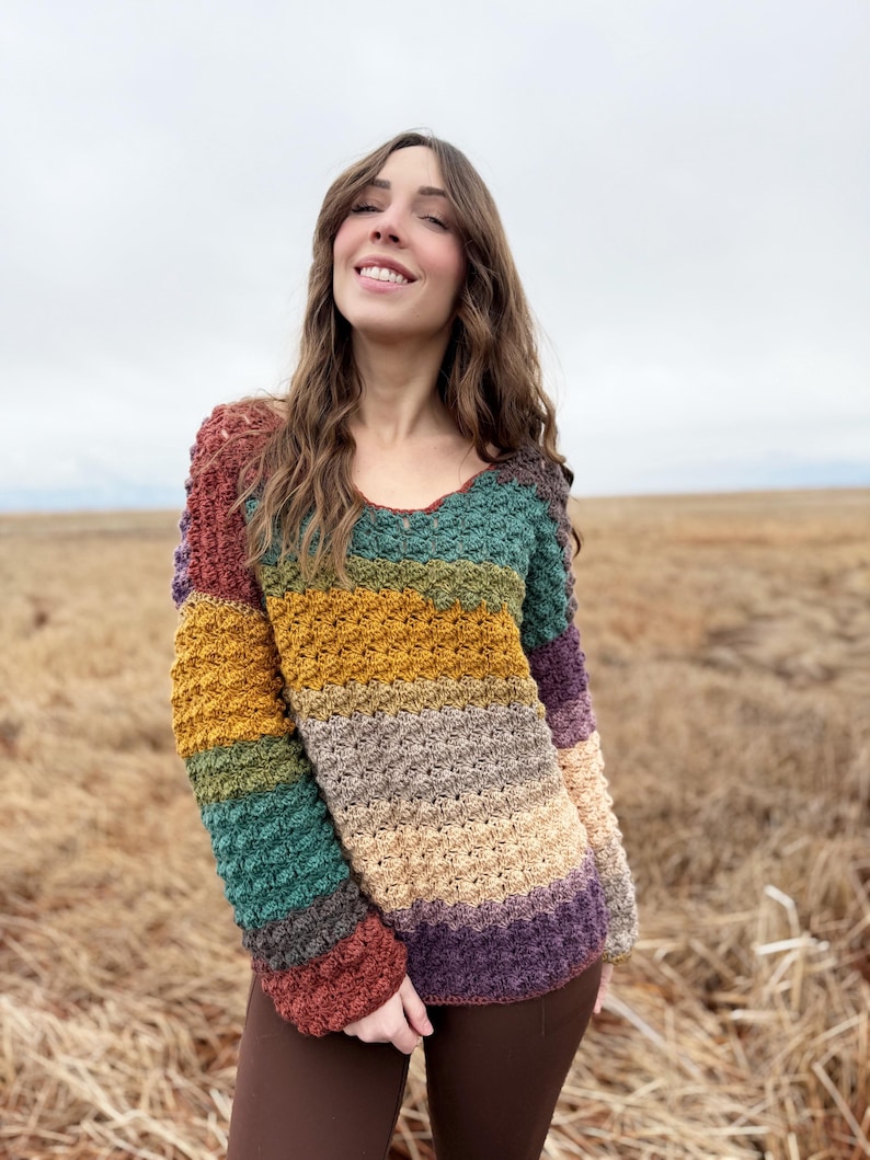 May include: A multi-coloured striped crochet sweater with long sleeves. The sweater is made with a textured stitch and features a variety of colours, including rust, teal, mustard yellow, beige, and purple.