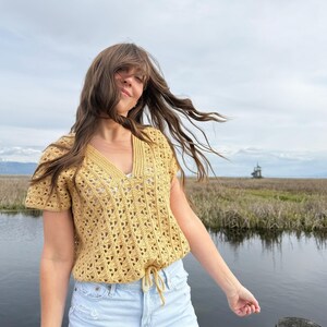May include: Mustard yellow crochet top with a V-neck and short sleeves. The top has a drawstring at the waist and a textured openwork design. The model is wearing light blue denim shorts. The background includes a body of water and a cloudy sky.
