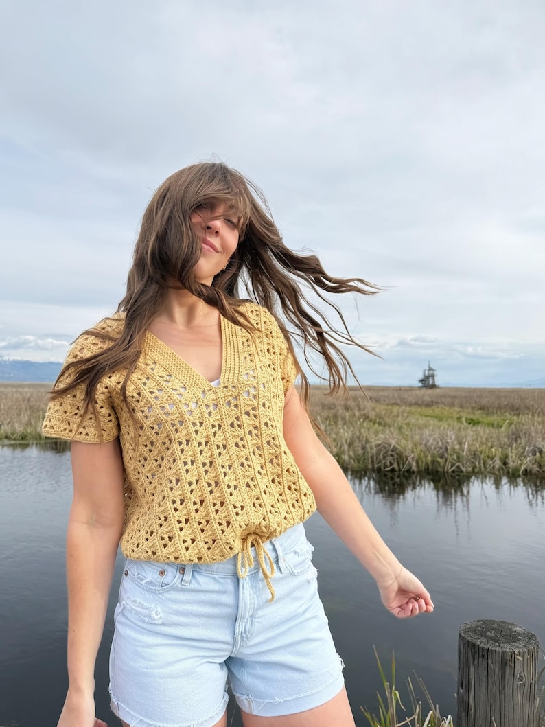 May include: Mustard yellow crochet top with a V-neck and short sleeves. The top has a drawstring at the waist and a textured openwork design. The model is wearing light blue denim shorts. The background includes a body of water and a cloudy sky.