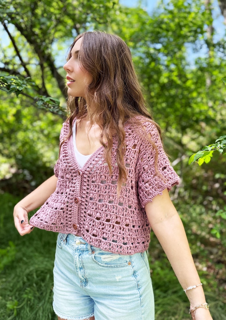 May include: A dusty rose crochet cardigan with short sleeves and a button-up front. The open weave design reveals a white undershirt. The cardigan is paired with light blue denim shorts. The background is a blurred view of green foliage.