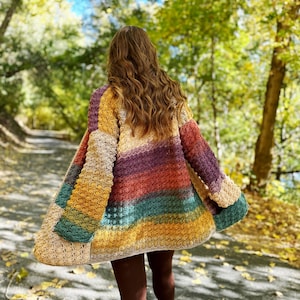 May include: A woman wearing a colourful striped crochet cardigan with a loose fit and long sleeves. The cardigan is made with a variety of colours, including mustard yellow, teal, burnt orange, and purple. The woman is walking on a path in a forest with autumn foliage.