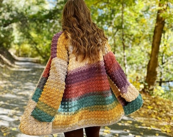 May include: A woman wearing a colorful striped crochet cardigan with a loose fit and long sleeves. The cardigan is made with a variety of colors, including mustard yellow, teal, burnt orange, and purple. The woman is walking on a path in a forest with fall foliage.
