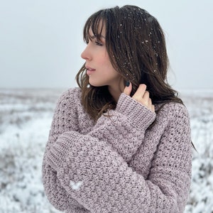 May include: A woman wearing a light purple crocheted cardigan in a snowy landscape. The cardigan has a textured stitch pattern and is worn with the sleeves crossed over the chest.