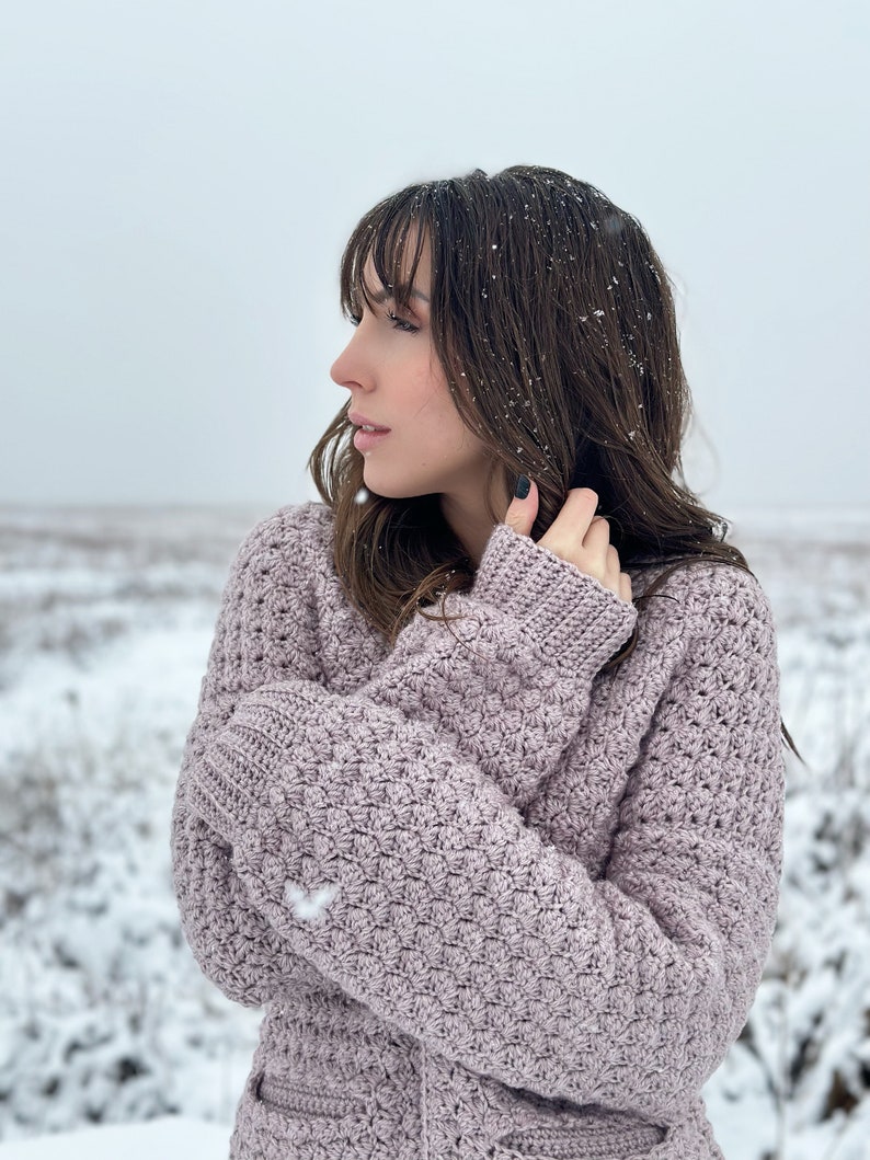 May include: A woman wearing a light purple crocheted cardigan in a snowy landscape. The cardigan has a textured stitch pattern and is worn with the sleeves crossed over the chest.