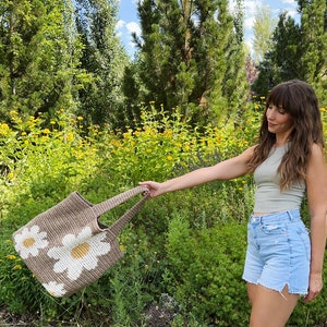 May include: A brown crocheted tote bag with white daisy flowers. The bag has two long straps and is being held by a person wearing a green tank top and blue denim shorts.