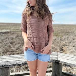 Puede incluir: Una mujer con un top de crochet marr&oacute;n con cuello en V y mangas cortas, pantalones cortos de mezclilla azul y zapatillas blancas. Est&aacute; de pie en una pasarela de madera con un banco de madera detr&aacute;s de ella.