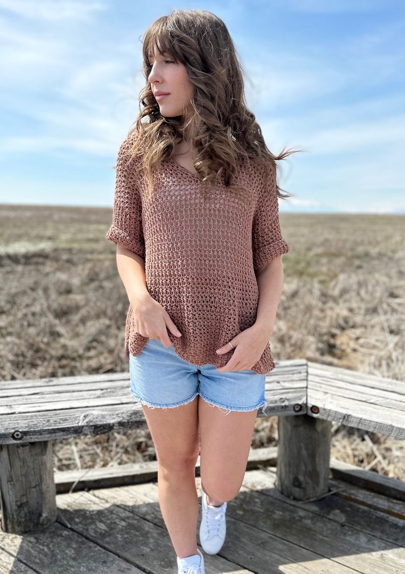 Puede incluir: Una mujer con un top de crochet marr&oacute;n con cuello en V y mangas cortas, pantalones cortos de mezclilla azul y zapatillas blancas. Est&aacute; de pie en una pasarela de madera con un banco de madera detr&aacute;s de ella.