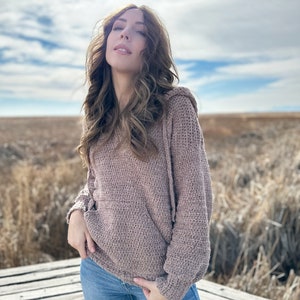 May include: A woman wearing a brown crocheted hooded sweater and blue jeans. She is standing outdoors in a field with a wooden platform behind her.