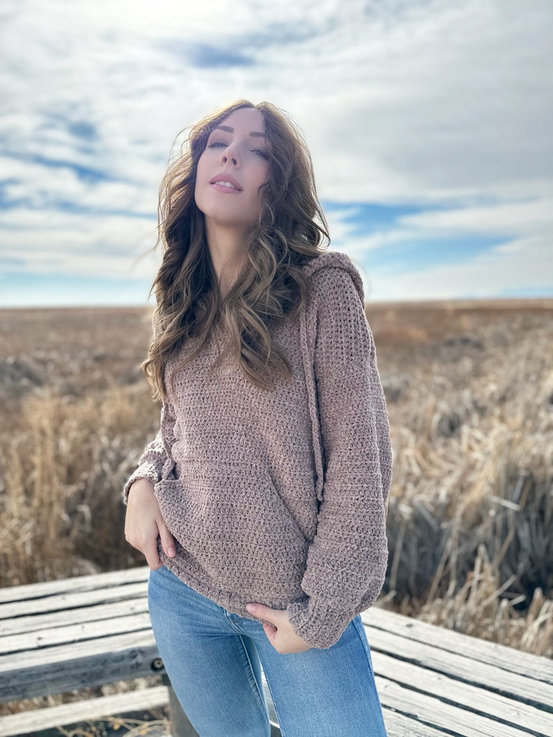 May include: A woman wearing a brown crocheted hooded sweater and blue jeans. She is standing outdoors in a field with a wooden platform behind her.