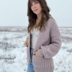 May include: A mauve crocheted cardigan with a textured stitch pattern and pockets. The cardigan is worn over a white tank top and blue jeans.
