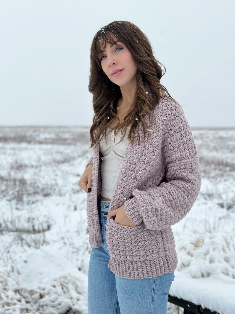 May include: A mauve crocheted cardigan with a textured stitch pattern and pockets. The cardigan is worn over a white tank top and blue jeans.
