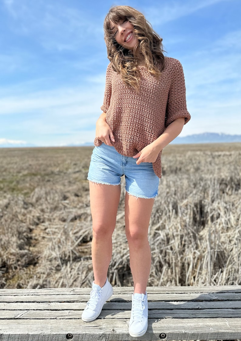 Puede incluir: Una mujer con un top de crochet marr&oacute;n, pantalones cortos de mezclilla azul claro y zapatillas blancas de ca&ntilde;a alta. Est&aacute; de pie en una plataforma de madera con un campo de hierba seca al fondo.