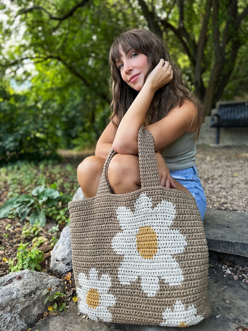 May include: A brown crocheted tote bag with white daisy flowers and yellow centres. The bag is large enough to carry groceries or other items.