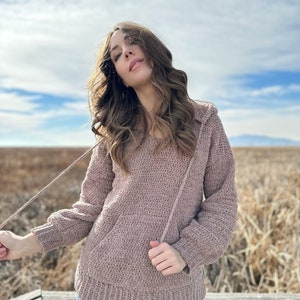 May include: A mauve crocheted hooded sweater with a relaxed fit and long sleeves. The sweater is worn by a person standing on a wooden platform.