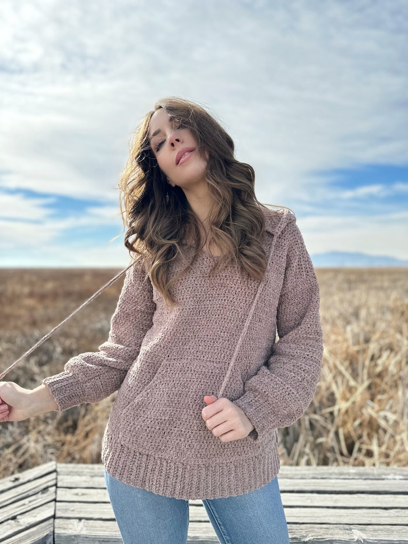 May include: A mauve crocheted hooded sweater with a relaxed fit and long sleeves. The sweater is worn by a person standing on a wooden platform.