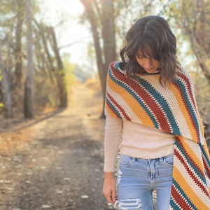 May include: A woman wearing a striped crocheted shawl in shades of rust, mustard yellow, teal, and cream. The shawl is draped diagonally across her shoulders and is worn with a cream-colored long-sleeved shirt and blue jeans.