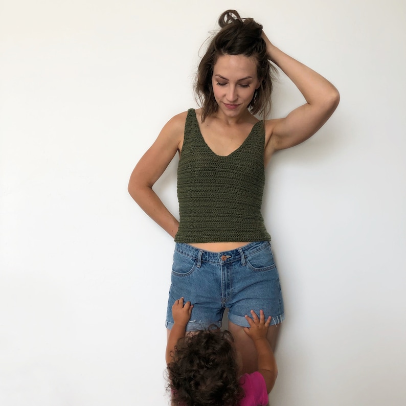 May include: A woman wearing a green crochet tank top and denim shorts. A young child is standing in front of her, reaching up towards her.