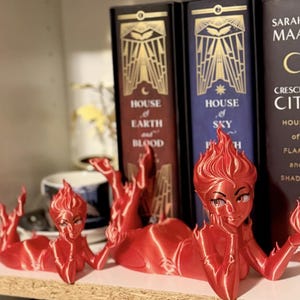 May include: Red, stylized female figure bookends with flame-like hair and outstretched arms. The bookends are positioned in front of a shelf with books, including titles like "House of Earth and Blood" and "Crescent City."
