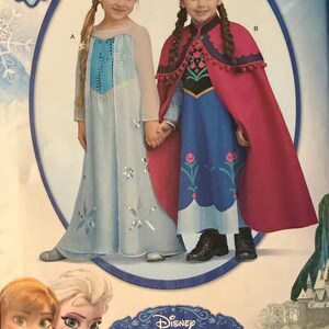 May include: A Disney Frozen home sewing pattern envelope labeled 1233 for sizes 3-8. The front displays two costumes: a light blue gown with silver snowflake accents and a dark blue dress paired with a magenta cape featuring pom-pom trim.