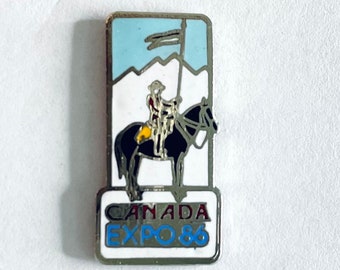 May include: A silver pin with a white background featuring a black and white illustration of a person on horseback holding a flag. The text "CANADA EXPO '86" is printed in red, blue, and white on the bottom of the pin.