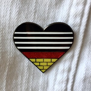 May include: A heart-shaped pin with a black and white striped design, a red glitter section, and a yellow brick pattern at the bottom.