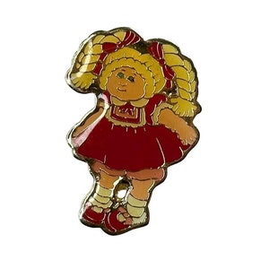 May include: A gold-colored pin featuring a cartoon image of a Cabbage Patch Kid doll with blonde pigtails, wearing a red dress with a white collar and a white bow. The doll has a smiling face with green eyes and a pink nose.