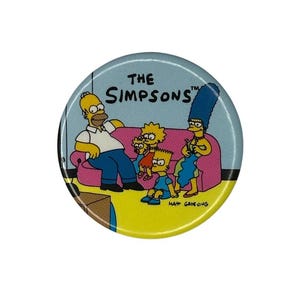May include: A yellow and blue button featuring the Simpsons family sitting on a pink couch. The text "The Simpsons" is written in yellow letters on a blue background. The button is round and has a white border.