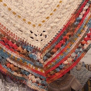May include: A white crocheted shawl with a colorful border. The border is made up of stripes of red, orange, yellow, blue, and green yarn. The shawl is draped over a white surface.