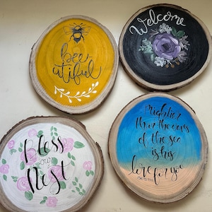 Könnte beinhalten: Vier handbemalte Holzscheiben mit dekorativen Designs. Eine zeigt eine Biene und die Worte "bee-utiful". Eine andere sagt "Welcome" mit einem Blumendesign. Die dritte sagt "Bless our Nest" mit Blumen, und die letzte hat ein Zitat.