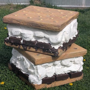 May include: Two-tiered, oversized s'mores sculpture. The graham cracker layers are light brown with small, round indentations. White marshmallow and dark chocolate bars are between the graham crackers. The sculpture is on green grass.