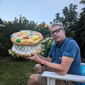 May include: A giant cookie ice cream sandwich with colourful chocolate sweets on top. The cookie is golden-brown, and the ice cream filling is white. The product is held by a person sitting outdoors.