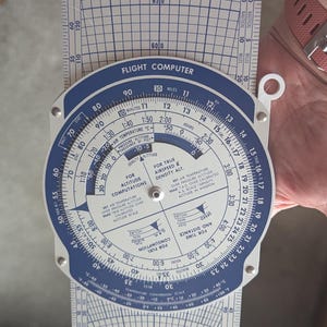 May include: A blue and white Piper Flight Computer PC-3. The circular slide rule has scales for altitude, airspeed, and density calculations. The device has a white handle and is designed for aviation use.