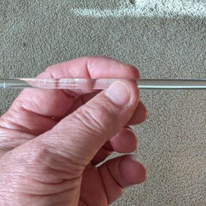 May include: A clear glass stirring rod with a slightly textured, ridged center section. The rod is held in a hand against a neutral-toned background. The rod appears to be approximately 15 cm long.