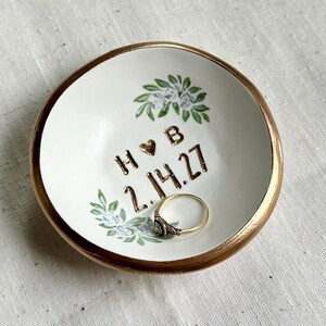 May include: A small, white ceramic ring dish with a gold rim. The dish is decorated with floral accents and the initials "H & B" with a heart symbol, and the date "2.14.27". A ring rests inside the dish.