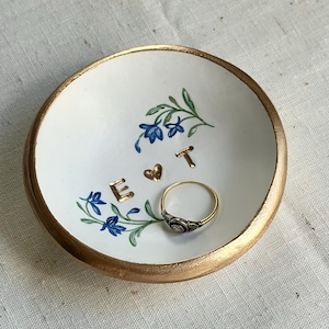 May include: A small, white ceramic ring dish with a gold rim. The dish features blue floral accents and the gold letters "E & T". A gold ring with a small stone sits inside the dish.