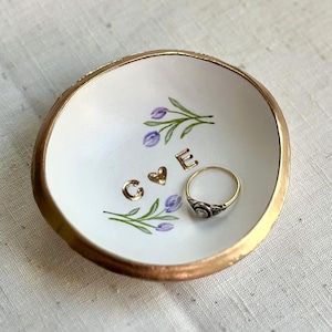 May include: A small, white ceramic ring dish with a gold rim and a gold-colored base. The dish features hand-painted purple flowers and the gold letters "C & E". A gold ring with a dark stone sits inside the dish.