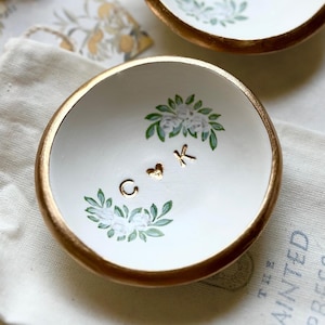 May include: Two small, white ceramic bowls with gold rims. Each bowl features a floral design with green leaves and white flowers, and gold initials. The bowls are on a white surface with lace and a small bag.