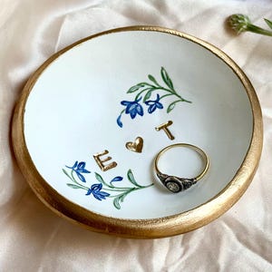 May include: A round white ceramic trinket dish with a gold-painted rim. The center features hand-painted blue cornflowers, the gold-colored letters E and T, and a small gold heart. A silver ring with a clear stone rests inside the dish.