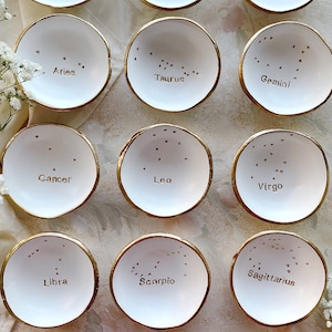 Puede incluir: Doce platos de anillo de cer&aacute;mica blanca con bordes dorados, cada uno con una constelaci&oacute;n del zod&iacute;aco diferente y su nombre. Las constelaciones son: Capricornio, Acuario, Piscis, Aries, Tauro, G&eacute;minis, C&aacute;ncer, Leo, Virgo, Libra, Escorpio y Sagitario.