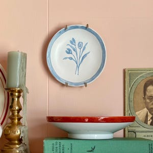 May include: A shelf displays a collection of decorative items. A white plate with a blue floral design hangs on the wall. A gold candlestick holds a gray candle. A red and white bowl sits atop books, and a vintage book with a portrait is visible.