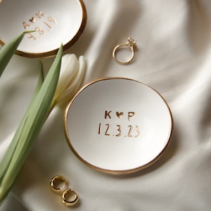 May include: Two white ceramic ring dishes with gold rims and personalised text. One dish has "A K 4.8.19" and the other has "K ♡ P 12.3.23". A gold ring and earrings are also present, with a white tulip and green leaves.