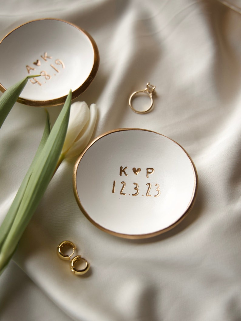 May include: Two white ceramic ring dishes with gold rims and personalised text. One dish has "A K 4.8.19" and the other has "K ♡ P 12.3.23". A gold ring and earrings are also present, with a white tulip and green leaves.