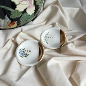 May include: Two small white ceramic ring dishes with gold accents. One dish has a floral design and the initials "E & M" written in gold. The other dish has a leafy design and the initials "A & E" written in gold.