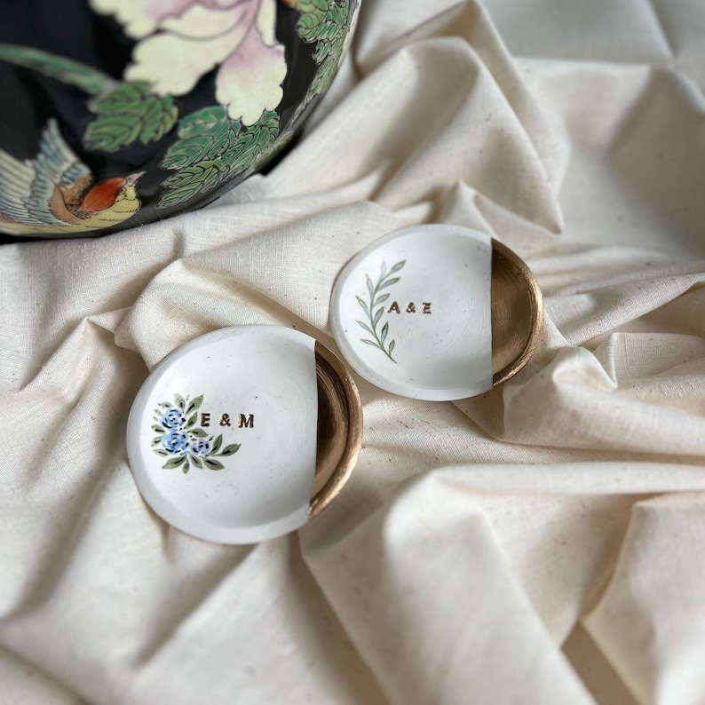 May include: Two small white ceramic ring dishes with gold accents. One dish has a floral design and the initials "E & M" written in gold. The other dish has a leafy design and the initials "A & E" written in gold.