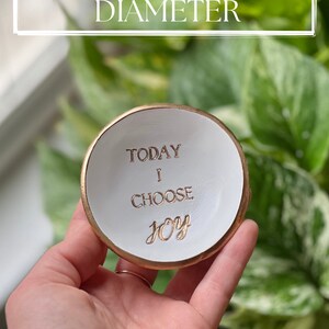 K&ouml;nnte beinhalten: Eine wei&szlig;e Keramik-Ring Schale mit goldfarbenem Rand. Die Schale hat einen Durchmesser von etwa 7,6 cm und tr&auml;gt die Aufschrift "Today I Choose Joy" in goldenen Buchstaben.