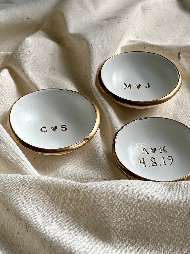 May include: Three white ceramic ring dishes with gold rims. Each dish has a different inscription: "C&hearts;S", "M&hearts;J", and "A&hearts;K 4.8.19".